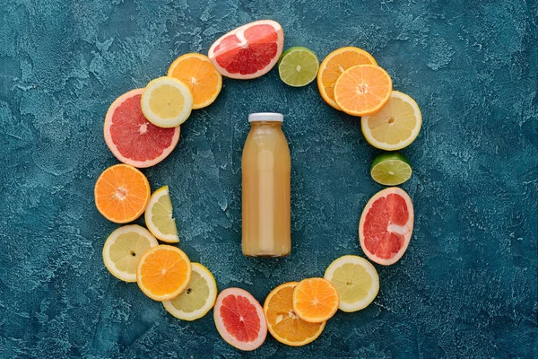 Top view of bottle of fresh juice surrounded with ripe citrus fruits slices in circle shape on blue concrete surface — Stock Photo
