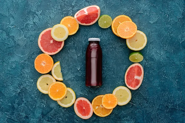 Vue du dessus de la bouteille de jus frais entourée de tranches d'agrumes en forme de cercle sur une surface en béton bleu — Photo de stock