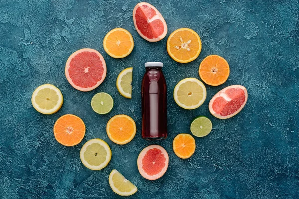 Top view of bottle of red juice surrounded with citrus fruits slices on blue concrete surface — Stock Photo