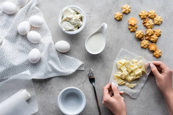 Vista parziale della donna che tiene il burro tagliato per torta con ingredienti disposti intorno sul tavolo grigio — Foto stock