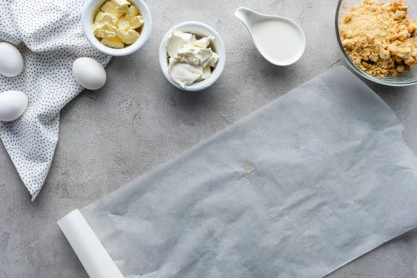 La puesta plana con papel de hornear, galletas trituradas, huevos de pollo crudos y mantequilla para hacer pastel en la superficie gris - foto de stock