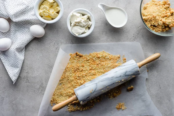 Flat lay with rolling pin, crushed cookies, raw chicken eggs and butter for making pie on grey surface — Stock Photo