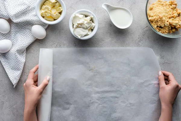 Tiro recortado de mujer sosteniendo papel de hornear con ingredientes arreglados para pastel en la mesa gris - foto de stock