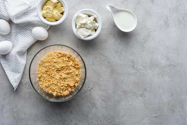 Flache Lage mit zerdrückten Plätzchen, rohen Hühnereiern und Butter für die Zubereitung von Kuchen auf grauer Oberfläche — Stockfoto