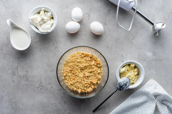 Flache Lage mit zerdrückten Plätzchen, rohen Hühnereiern und Butter für die Zubereitung von Kuchen auf grauer Oberfläche — Stockfoto