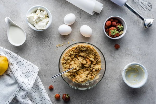 Vista dall'alto della pasta preparata per torta, fragole, uova di pollo crude e carta da forno su tavolo grigio — Foto stock