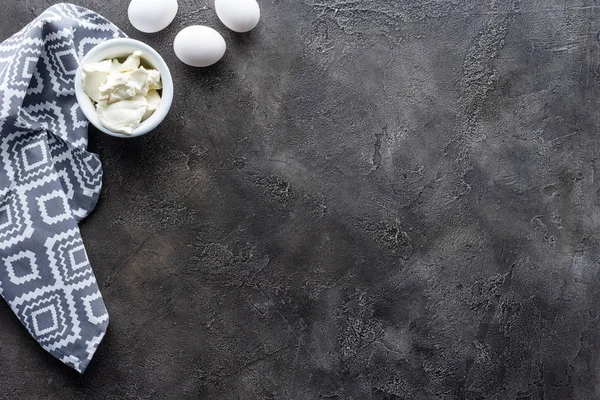 Vista superior de los huevos de pollo crudos arreglados, crema agria en un tazón y ropa de cama sobre una mesa gris oscuro - foto de stock