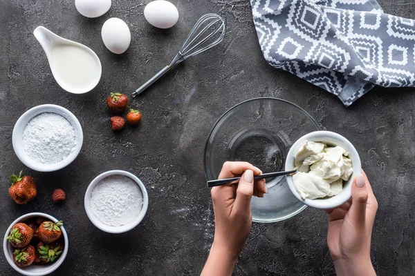 Vista parcial de la mujer poniendo crema agria en un tazón mientras hace crema para pastel en la superficie gris oscura - foto de stock