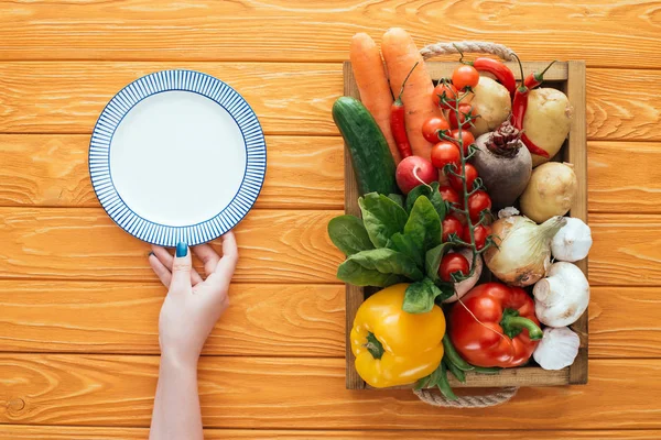 Обрезанный снимок человека, держащего пустую круглую тарелку и свежие сырые овощи в коробке на деревянном столе — стоковое фото