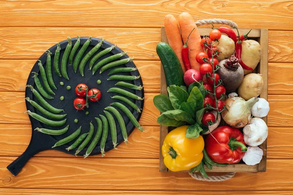Fresh green peas and tomatoes on round board and raw healthy vegetables in box on wooden table top — Stock Photo