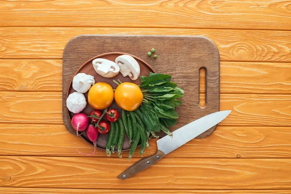 Top view of fresh raw vegetables in plate on wooden cutting board and knife on wooden table top — Stock Photo