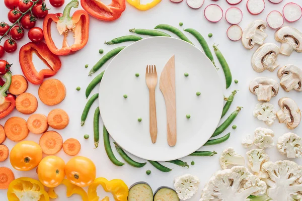 Vista superior de tenedor de madera y cuchillo en plato redondo blanco y verduras orgánicas frescas aisladas en blanco - foto de stock