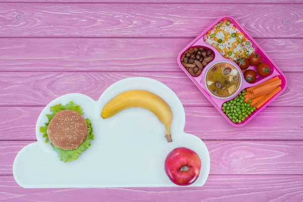 Vista superior de la bandeja con almuerzo para niños para la escuela, hamburguesa y frutas en la mesa rosa - foto de stock