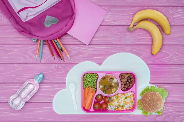 Вид зверху лоток з дитячим обідом для школи, рожевий мішок, олівці та банани на рожевому столі — стокове фото