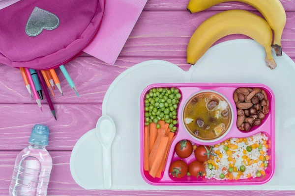 Вид зверху лоток з дитячим обідом для школи, рожевий мішок, олівці та пляшка води на рожевому столі — стокове фото