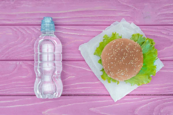 Vista superior de hamburguesa y botella de agua de plástico sobre mesa de madera violeta - foto de stock