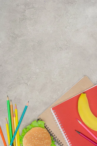 Top view of burger, banana, notebooks and pencils on marble table — Stock Photo