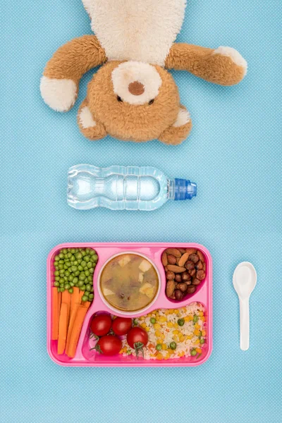 Вид зверху лоток з дітьми обід для школи, плюшевий ведмідь і пляшка води на синій поверхні — стокове фото