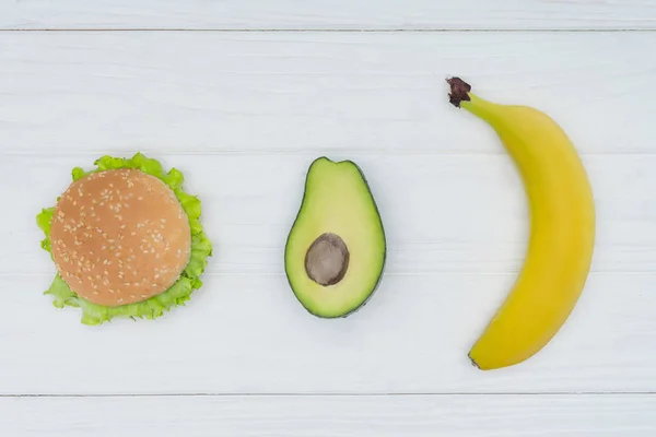 Draufsicht auf Burger, Avocado und Banane auf weißem Holztisch — Stockfoto