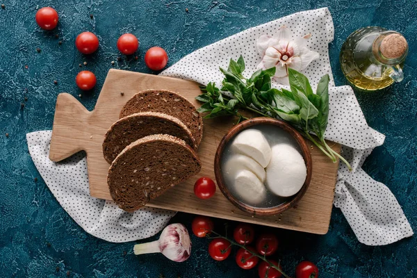 Tablero de madera con mozzarella y pan sobre mesa azul oscuro con tomates rojos - foto de stock