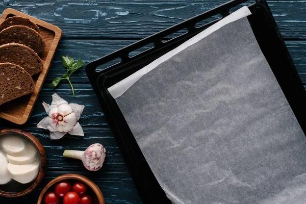 Empty baking sheet on dark wooden table by cooking ingredients and tomatoes — Stock Photo