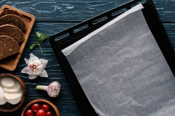 Hoja de hornear vacía en la mesa de madera oscura por ingredientes de cocina y tomates - foto de stock