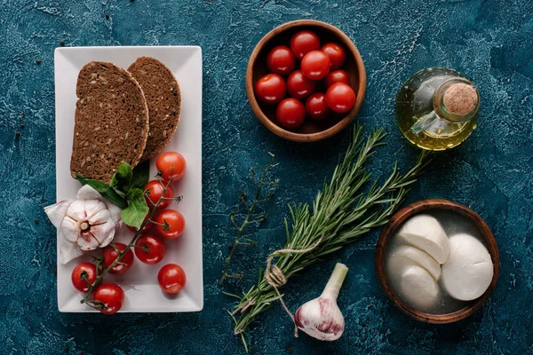 Mozzarella cheese and tomatoes on dark blue table with bread and herbs — Stock Photo
