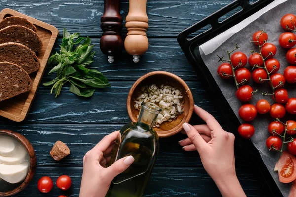 Vista recortada de las manos femeninas añadiendo aceite al ajo en la mesa de madera oscura con tomates y hierbas - foto de stock