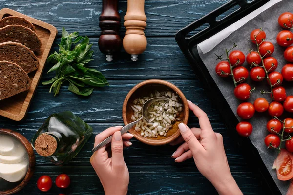 Vista recortada de manos femeninas cocinando aceite de ajo sobre mesa de madera oscura con tomates y hierbas - foto de stock