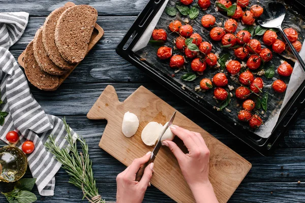 Frau schneidet Mozzarella für Sandwiches mit Kräutern und gebackenen Tomaten auf dunklem Holztisch — Stockfoto