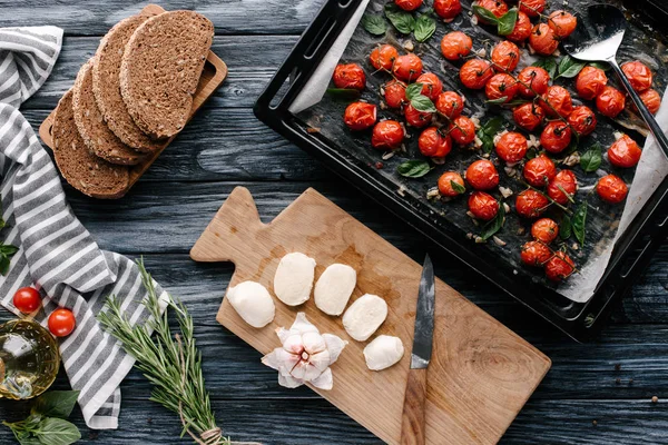 Кастрюля с запечёнными помидорами на тёмном деревянном столе с хлебом и сыром — стоковое фото