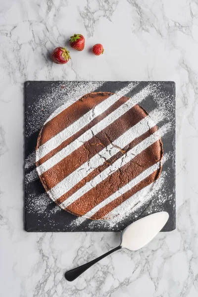 Tasty homemade brown cake with icing on slate board and fresh strawberries on marble surface — Stock Photo