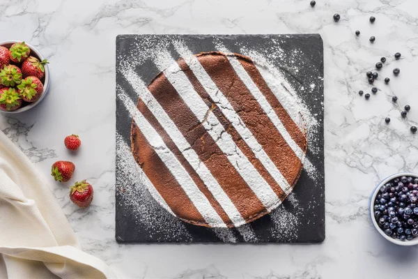 Draufsicht auf köstlichen hausgemachten braunen Kuchen mit Sahnehäubchen auf Schiefertafel und frischen Beeren auf Marmoroberfläche — Stockfoto