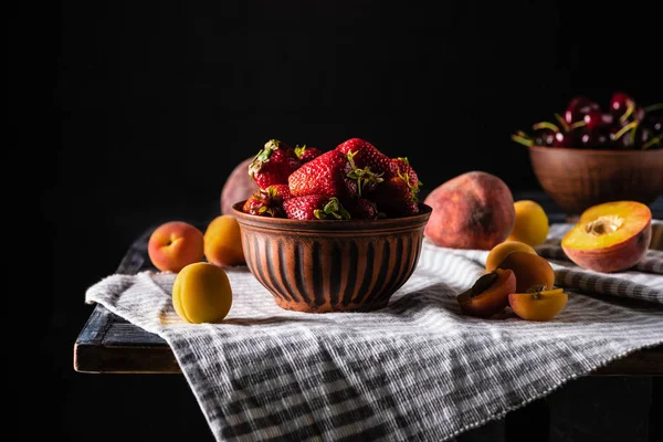 Enfoque selectivo de tazón con fresas rodeadas de melocotones y albaricoques en la mesa de madera en negro - foto de stock