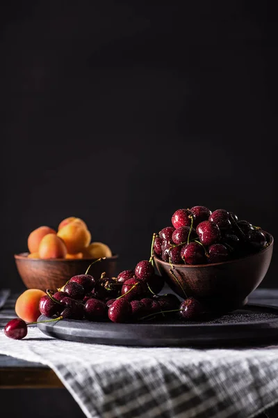 Selektiver Fokus von Schalen mit reifen Kirschen und Aprikosen auf Holztisch auf schwarz — Stockfoto