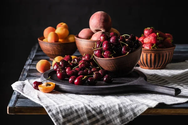 Foco selectivo de cuencos con cerezas, fresas, melocotones y albaricoques en mesa de madera en negro - foto de stock