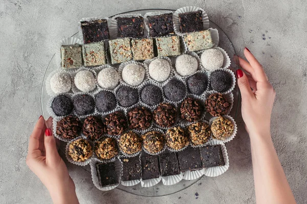 Vista parcial de la mujer sosteniendo el plato con varios postres dulces en la mesa gris - foto de stock