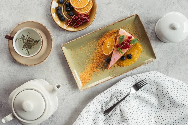 Acostado plano con trozo de pastel de zanahoria dulce con relleno de bayas, tetera y taza de té de hierbas en la mesa gris - foto de stock