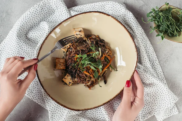 Visão parcial de mãos femininas e soba com tofu e legumes decorados com sementes germinadas de girassol em mesa cinza — Fotografia de Stock