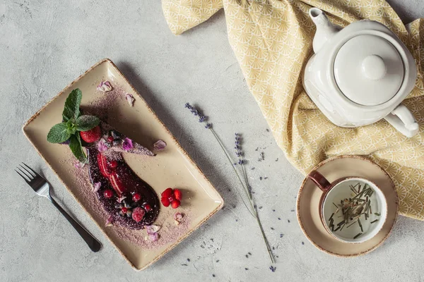 Composition alimentaire avec morceau de gâteau aux myrtilles servi avec des feuilles de menthe et des pétales violets sur assiette, théière et tasse de tisane sur plateau gris — Photo de stock