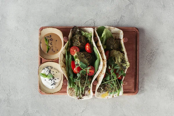 Top view of falafel with tortillas, cherry tomatoes and germinated seeds of sunflower served on wooden board on grey surface — Stock Photo