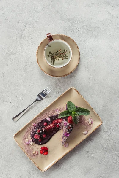 Essenszusammensetzung mit einem Stück Heidelbeerkuchen, serviert mit Minzblättern und Veilchenblättern auf dem Teller und einer Tasse Kräutertee auf der grauen Tischplatte — Stockfoto