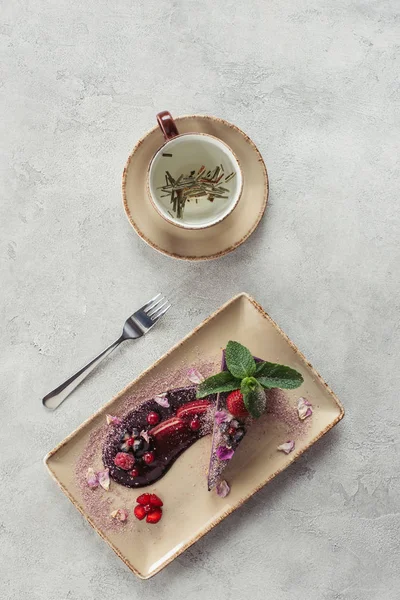 Composition alimentaire avec morceau de gâteau aux myrtilles servi avec des feuilles de menthe et des pétales violets sur assiette et tasse de tisane sur plateau gris — Photo de stock