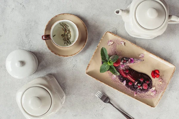 Food composition with piece of blueberry cake served with mint leaves and violet petals on plate, teapot and cup of herbal tea on grey tabletop — Stock Photo