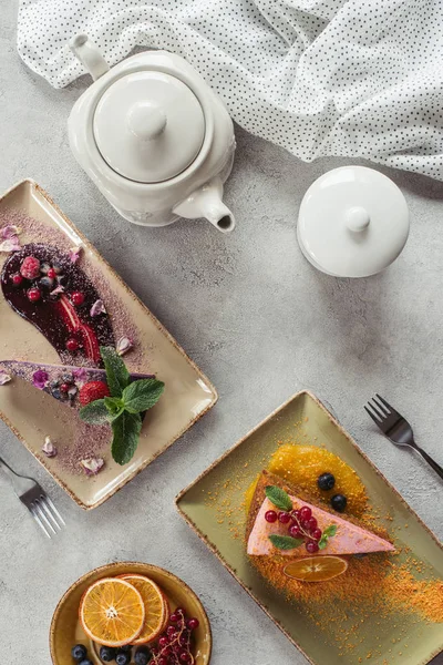 Flat lay com bolo de cenoura doce com recheio de baga, bolo de mirtilo servido com folhas de hortelã e pétalas violetas, bule e linho em mesa cinza — Fotografia de Stock