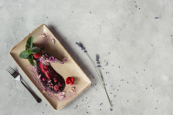 Пищевой состав с кусочком черничного торта подается с мятными листьями и фиолетовыми лепестками на тарелке на сером столе — стоковое фото