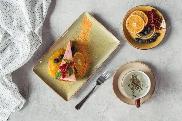Flache Lage mit süßem Karottenkuchen mit Beerenfüllung, serviert mit Orangenscheibe, Tasse Kräutertee und Gabel auf grauer Tischplatte — Stockfoto