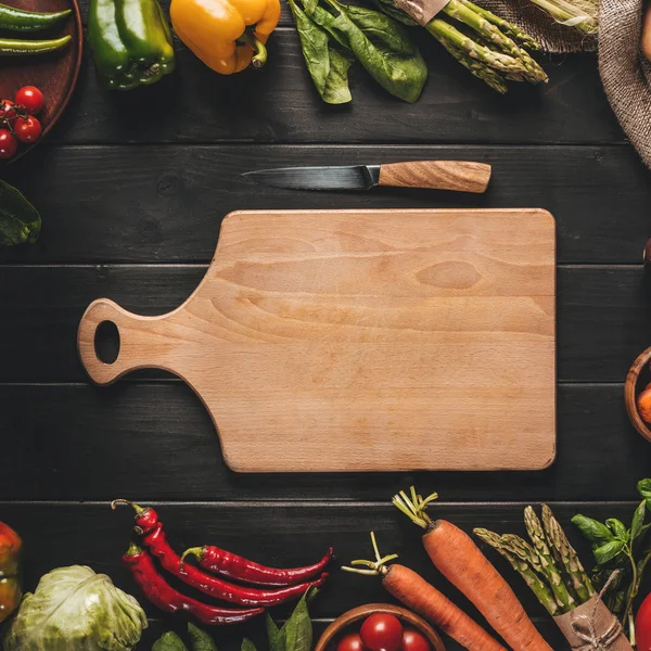 Draufsicht auf Holzschneidebrett mit Messer und frischem Bio-Gemüse auf schwarzer Holztischplatte — Stockfoto
