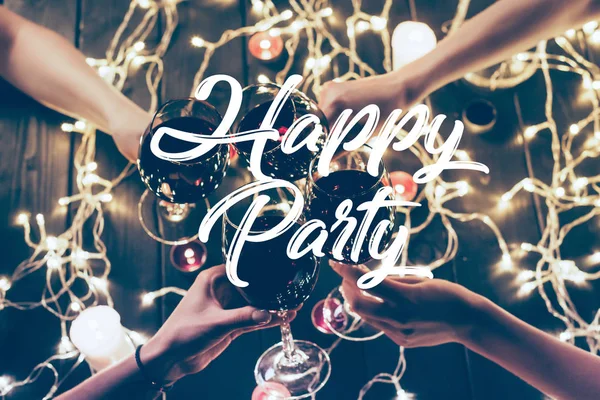 Cropped shot of four people clinking glasses with red wine over wooden table with fairylights decorations, happy party inscription — Stock Photo