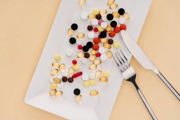 Вид сверху на тарелку с различными таблетками и вилкой с ножом на бежевом фоне — стоковое фото